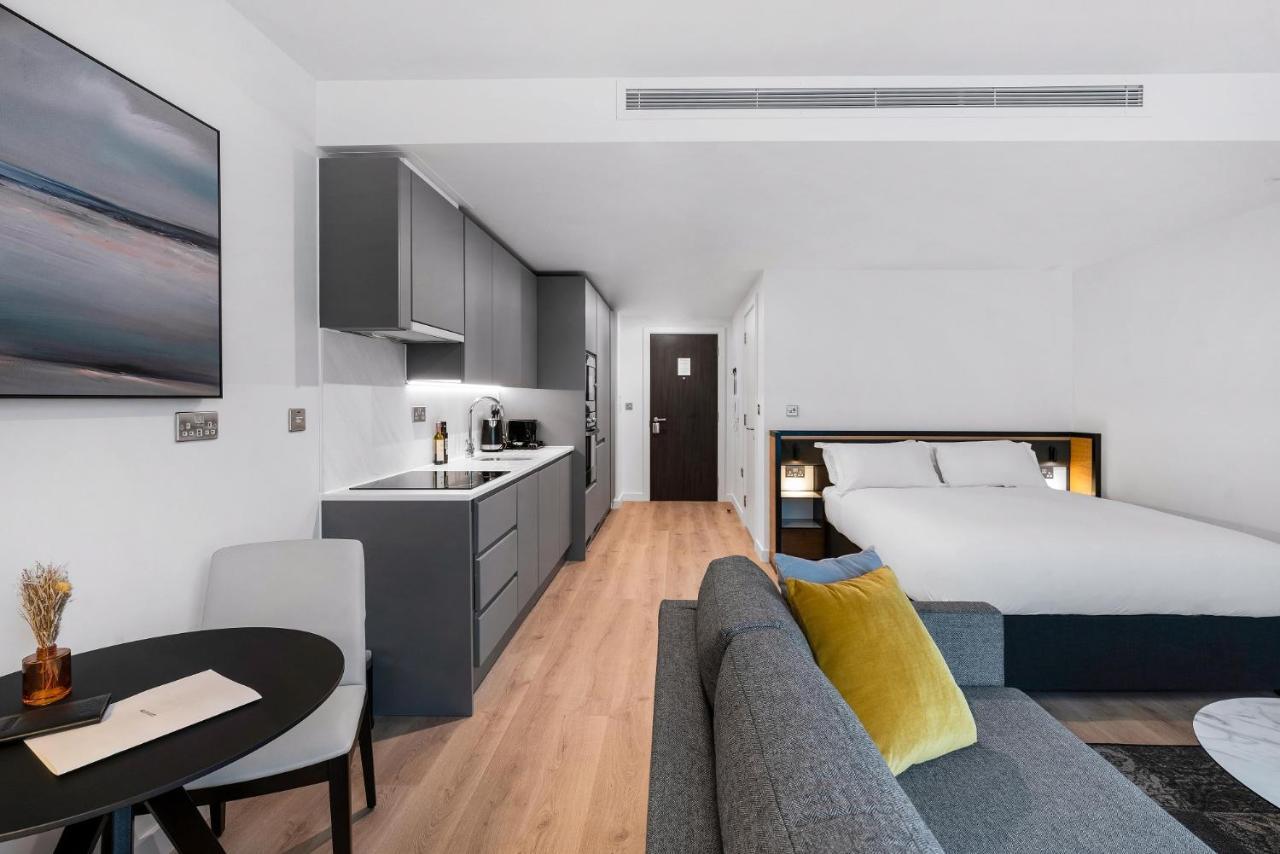 Citysuites 2 Aparthotel Manchester Exterior photo The photo shows a modern and stylish studio apartment. It features a kitchenette with dark cabinetry and a small dining table with two chairs. There's a cozy sofa with colorful cushions, positioned next to the table. In the background, a large bed wi