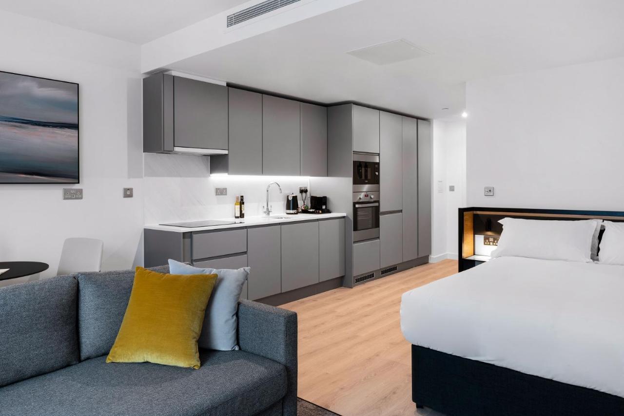 Citysuites 2 Aparthotel Manchester Exterior photo The photo shows a modern studio apartment featuring an open-plan layout. On one side, there is a kitchenette with sleek, gray cabinets and appliances, including a coffee maker and oven. Adjacent to the kitchenette is a small dining area with a table 