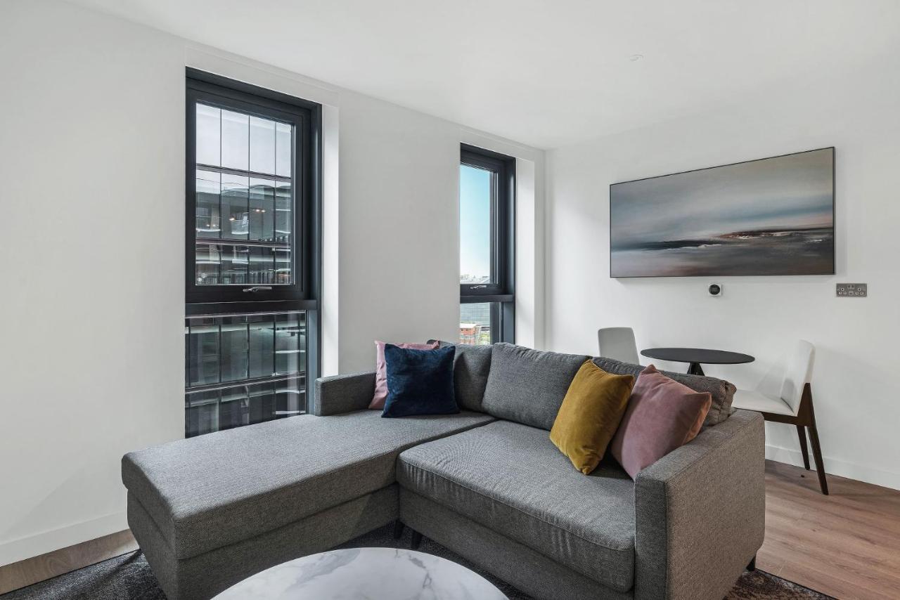 Citysuites 2 Aparthotel Manchester Exterior photo The photo shows a modern living space featuring a gray sectional sofa adorned with colorful throw pillows. There are large windows allowing natural light to fill the room, offering a view of the outdoors. A wall-mounted television is positioned above