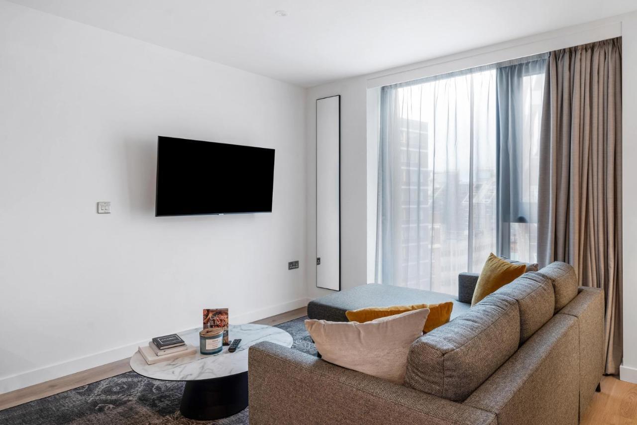 Citysuites 2 Aparthotel Manchester Exterior photo The image shows a modern living room with a minimalist design. There is a large flat-screen TV mounted on the white wall. A comfortable gray sofa with textured cushions occupies the center of the room, complemented by a round coffee table in front of