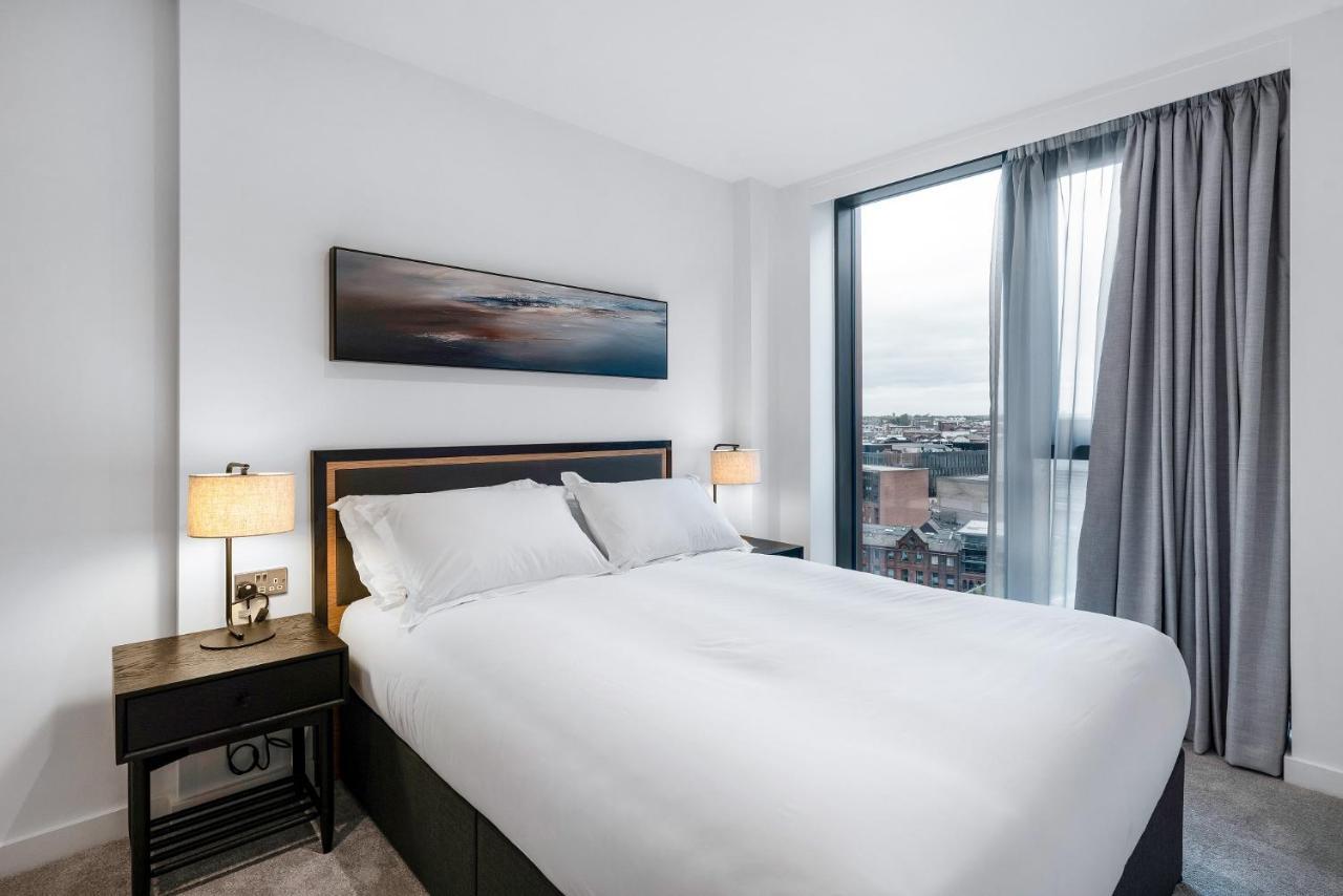 Citysuites 2 Aparthotel Manchester Exterior photo The image shows a modern bedroom featuring a large bed with a white duvet and two pillows. On either side of the bed are small nightstands with lamps. A large window occupies one wall, draped with light gray curtains, allowing natural light to enter 