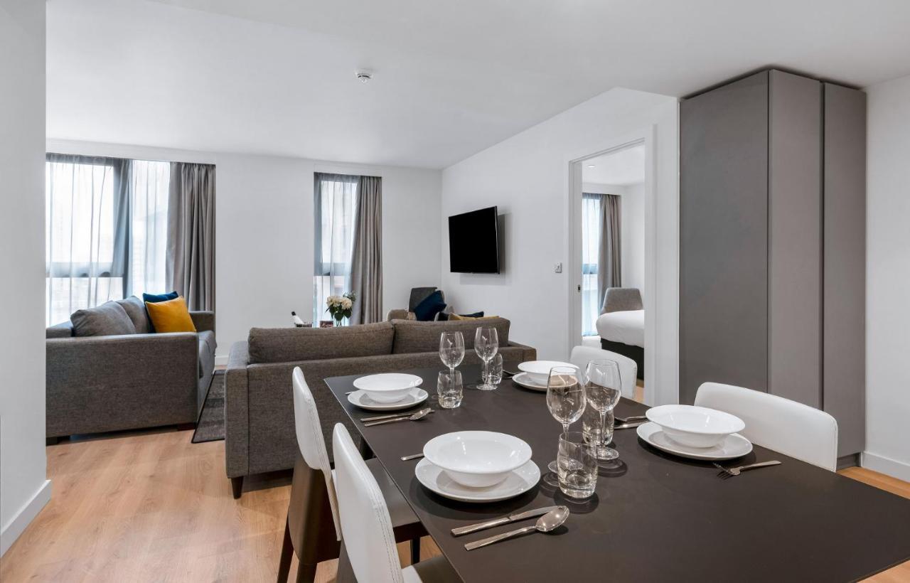 Citysuites 2 Aparthotel Manchester Exterior photo The photo shows a modern living space with an open layout. In the foreground, there is a dining table set for a meal, featuring multiple plates, glasses, and cutlery arranged neatly. In the background, there is a comfortable-looking gray sofa with a 