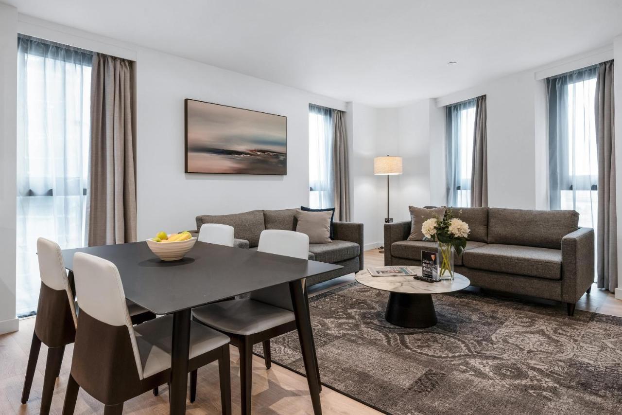 Citysuites 2 Aparthotel Manchester Exterior photo The photo depicts a modern and stylish interior of a living space. On the left, there is a dining table with four chairs, featuring a bowl of fruits on the table. In the background, there's a cozy seating area with a gray sofa and a smaller round cof