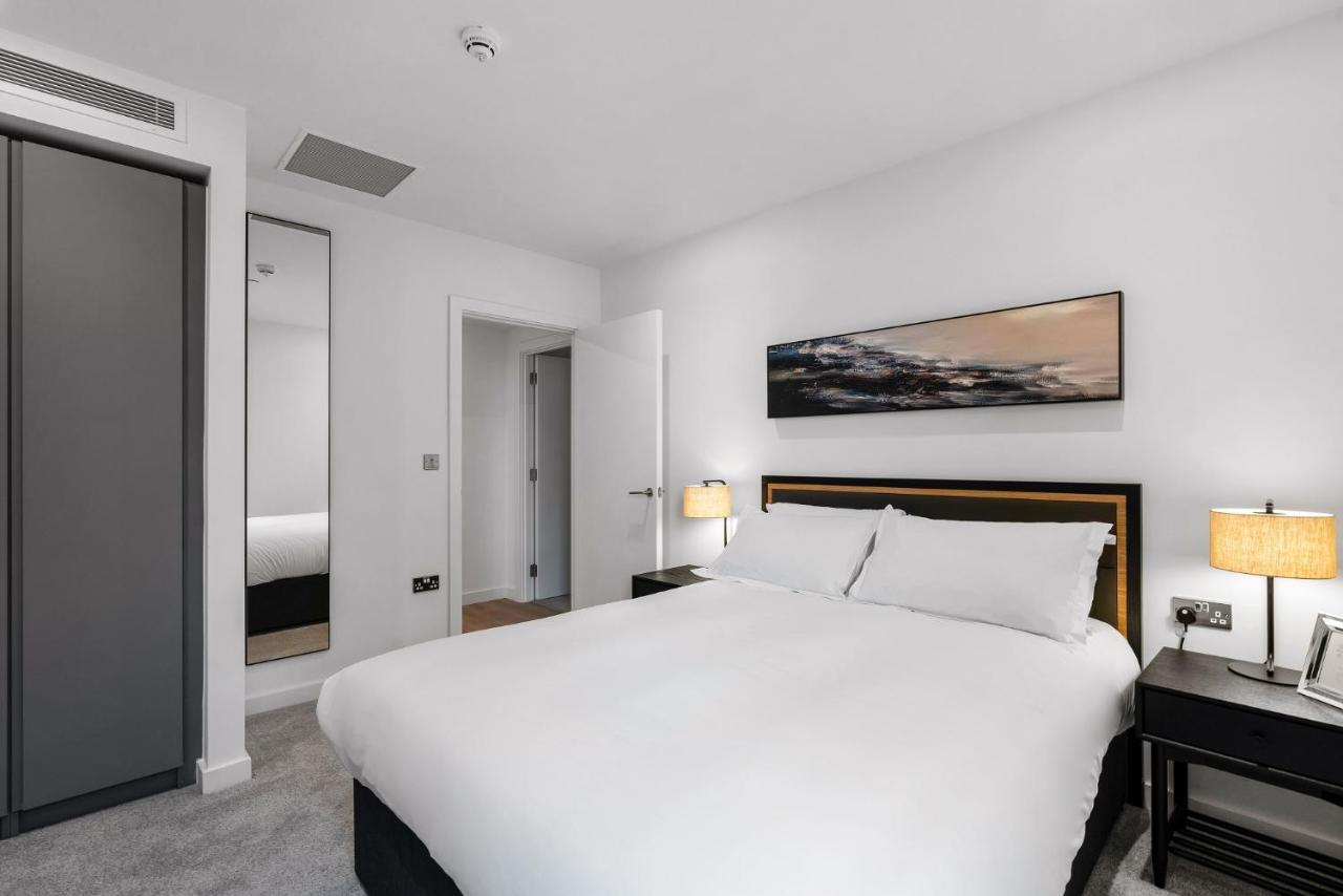 Citysuites 2 Aparthotel Manchester Exterior photo The photo shows a modern bedroom featuring a large double bed with white bedding. There are two bedside tables with lamps on either side of the bed. The walls are painted white, and there's a piece of artwork above the bed that depicts a landscape. A