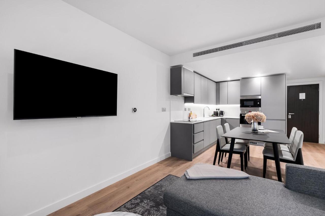 Citysuites 2 Aparthotel Manchester Exterior photo The photo shows a modern, minimalist living space. On the left, there is a large flat-screen TV mounted on the wall. To the right, a kitchen area features sleek gray cabinets and appliances, including a built-in microwave. In front of the kitchen, th