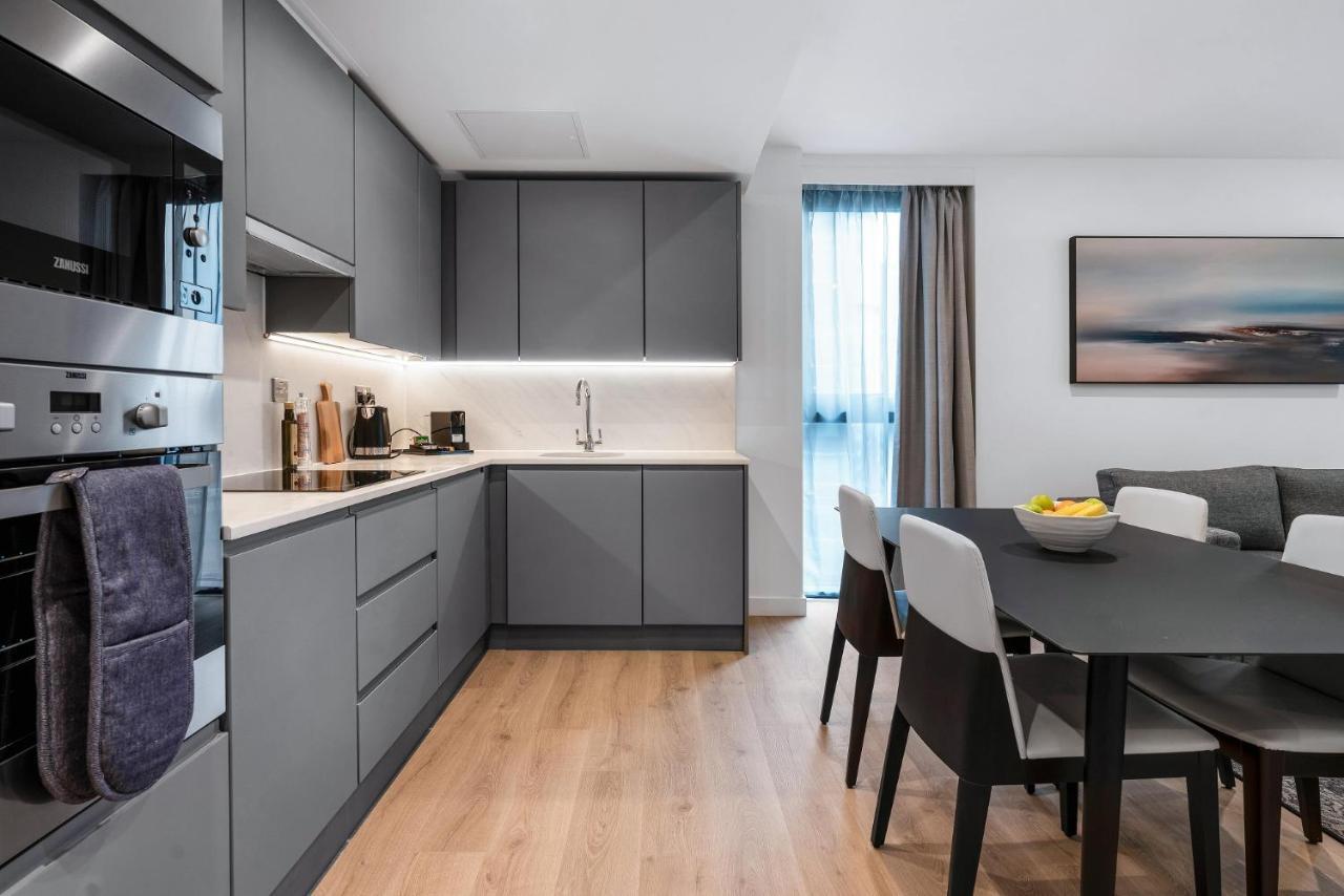Citysuites 2 Aparthotel Manchester Exterior photo The photo shows a modern kitchen and dining area. The kitchen features sleek gray cabinetry, a white countertop, and built-in appliances such as an oven. There is a sink and some kitchen utensils displayed. The adjacent dining area contains a black t