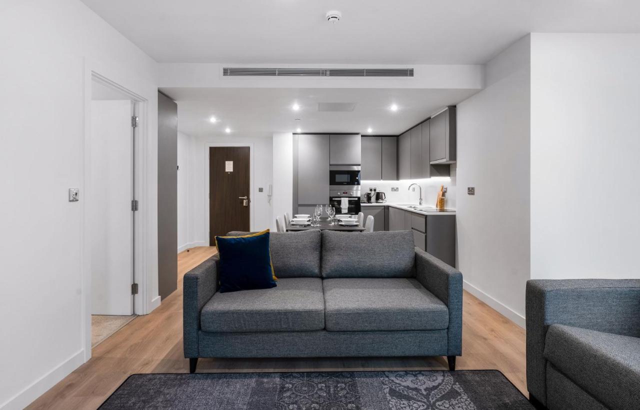 Citysuites 2 Aparthotel Manchester Exterior photo The photo shows a modern interior space, likely a living area or apartment. In the foreground, there is a gray sofa with a blue cushion positioned on a textured rug. To the back, there is an open kitchen with sleek cabinetry and appliances, visible a