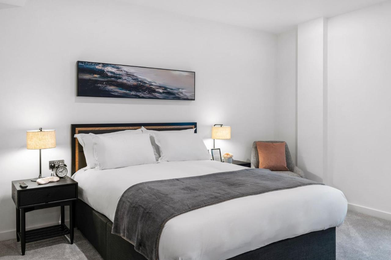 Citysuites 2 Aparthotel Manchester Exterior photo The photo shows a modern bedroom featuring a large bed with crisp white linens and a grey throw blanket. On either side of the bed, there are bedside tables with lamps. A piece of abstract art hangs above the headboard, and there is a chair in a corn