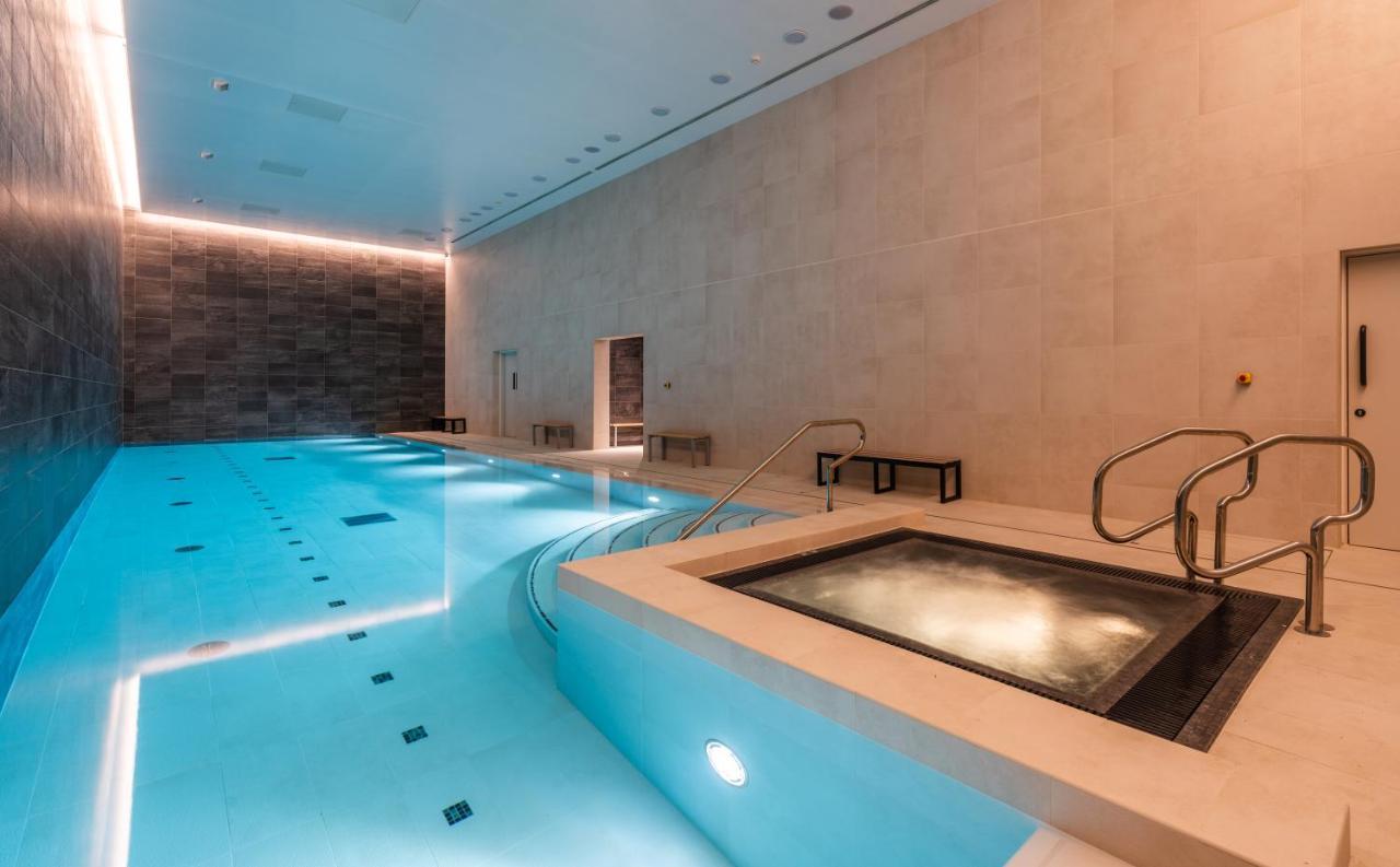 Citysuites 2 Aparthotel Manchester Exterior photo The photo shows an indoor swimming pool area. The pool is a bright blue color and features smooth, tiled surfaces. On one side, there is a hot tub area with steps leading into it. The walls are made of light-colored stone, and there is ambient lighti