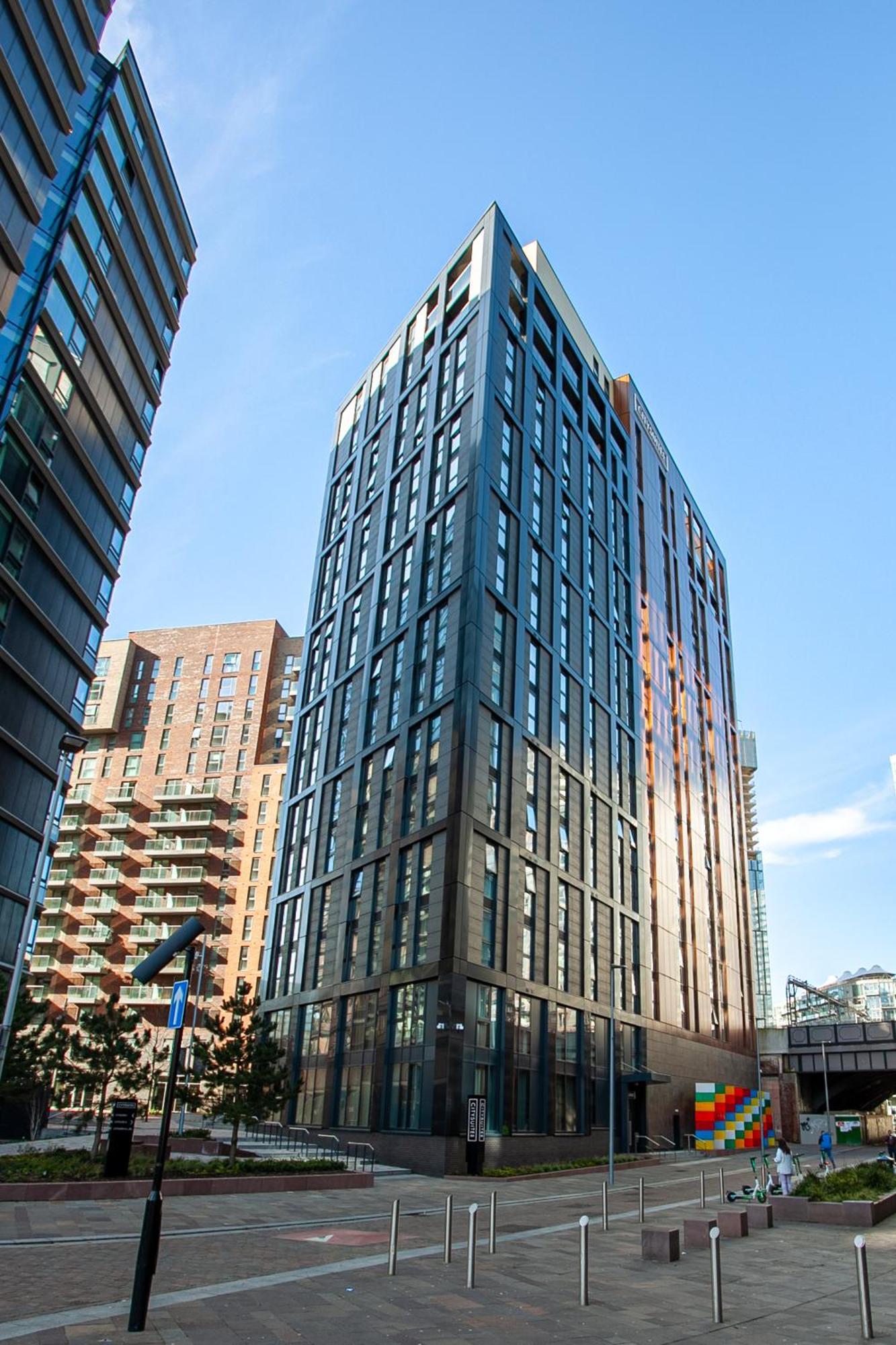 Citysuites 2 Aparthotel Manchester Exterior photo The photo shows a modern high-rise building with a sleek, glass façade. It is set against a blue sky, indicating a clear day. The building appears to have multiple floors with large windows that reflect light. Nearby, there are additional buildings, 